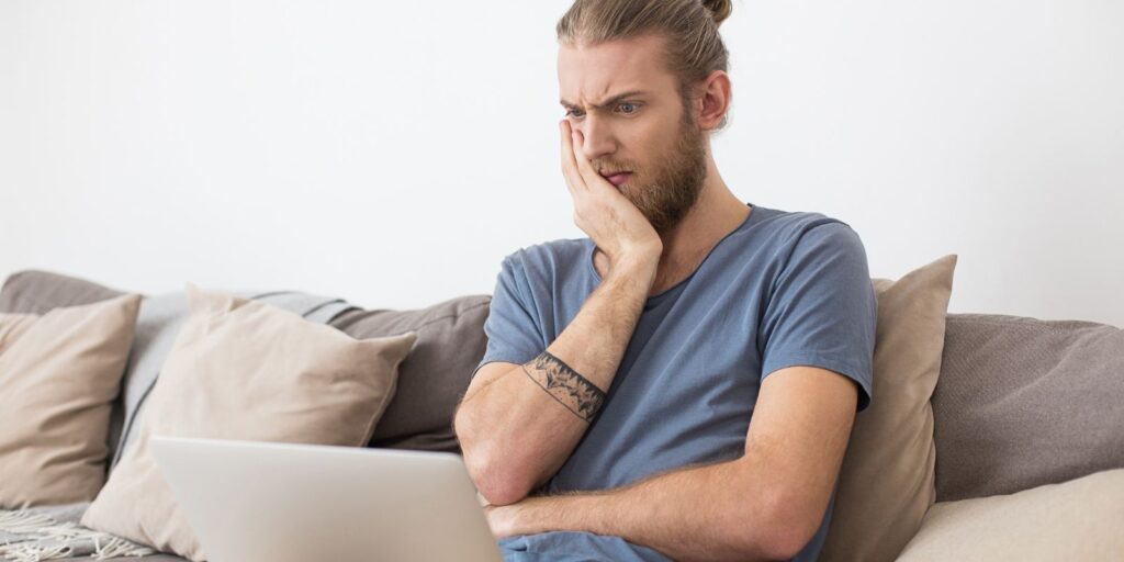Ritratto di giovane uomo seduto sul divano grigio che guarda stupito lo schermo computer portatile