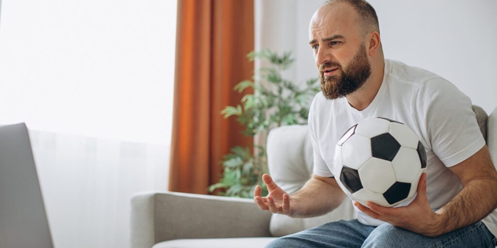 Uomo che tiene la palla da calcio e guarda il calcio online a casa – Scommettitore Professionista